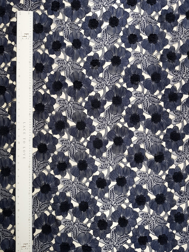 May include: A navy blue floral lace fabric featuring a repeating pattern of embroidered flowers and leaves. A white ruler is placed along the left side, showing a length of 92 centimetres. The lace has a delicate, openwork design with textured centres.