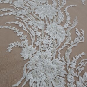 Puede incluir: Tela de encaje floral blanca con adornos de cuentas. El encaje presenta un delicado dise&ntilde;o floral con detalles intrincados y un brillo sutil.
