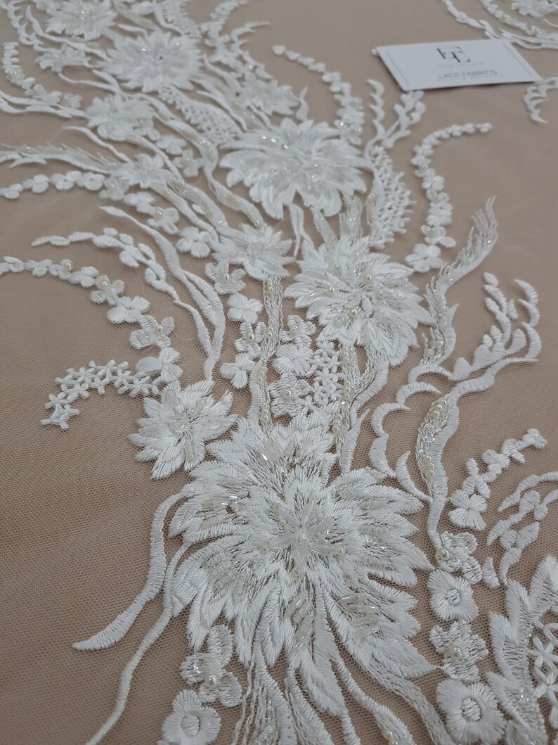 Puede incluir: Tela de encaje floral blanca con adornos de cuentas. El encaje presenta un delicado dise&ntilde;o floral con detalles intrincados y un brillo sutil.