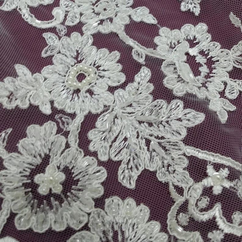 May include: Close-up of white floral lace with beaded accents. The lace features detailed flower and leaf patterns, with small pearls at the center of some flowers. The lace is set against a dark purple background.