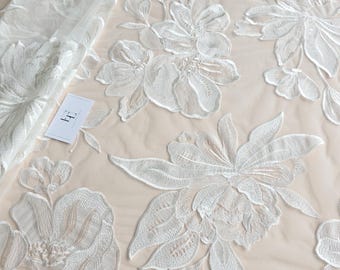 May include: White floral lace fabric with large, detailed embroidered flowers and leaves. The delicate design is set against a light beige background. The fabric is ideal for bridal wear, dresses, or other special occasion projects.