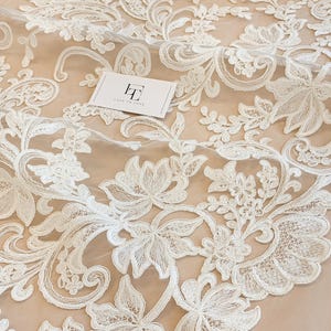 May include: White floral lace fabric featuring intricate embroidery on a sheer mesh base. The pattern consists of swirling vines, leaves, and blossoms. A small white rectangular card with the black logo text Lace to Love is placed on the fabric.