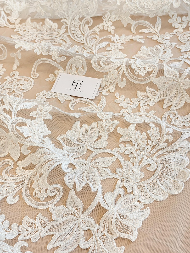 May include: White floral lace fabric featuring intricate embroidery on a sheer mesh base. The pattern consists of swirling vines, leaves, and blossoms. A small white rectangular card with the black logo text Lace to Love is placed on the fabric.