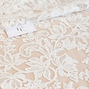 May include: White floral lace fabric featuring intricate embroidery and scalloped edges layered over a solid beige mesh base. A small white rectangular tag with the black logo LT and the text LACE TO LOVE is placed on the fabric surface.