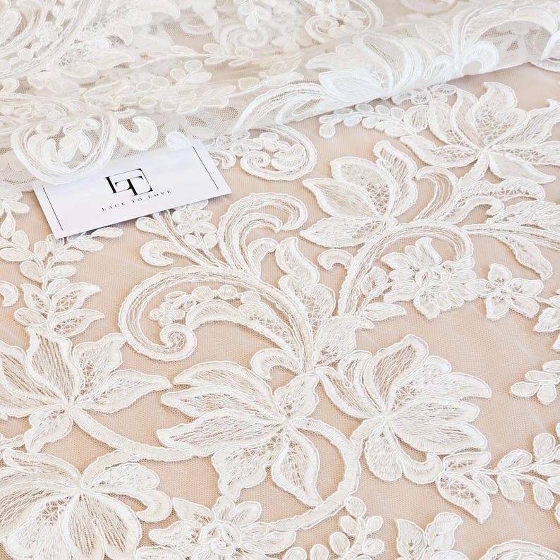 May include: White floral lace fabric featuring intricate embroidery and scalloped edges layered over a solid beige mesh base. A small white rectangular tag with the black logo LT and the text LACE TO LOVE is placed on the fabric surface.
