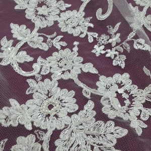May include: Close-up of white floral lace fabric with intricate embroidered details. The lace features a delicate pattern of flowers, leaves, and beaded accents. The fabric is set against a deep purple background, creating a contrast that highlights the lace's design.