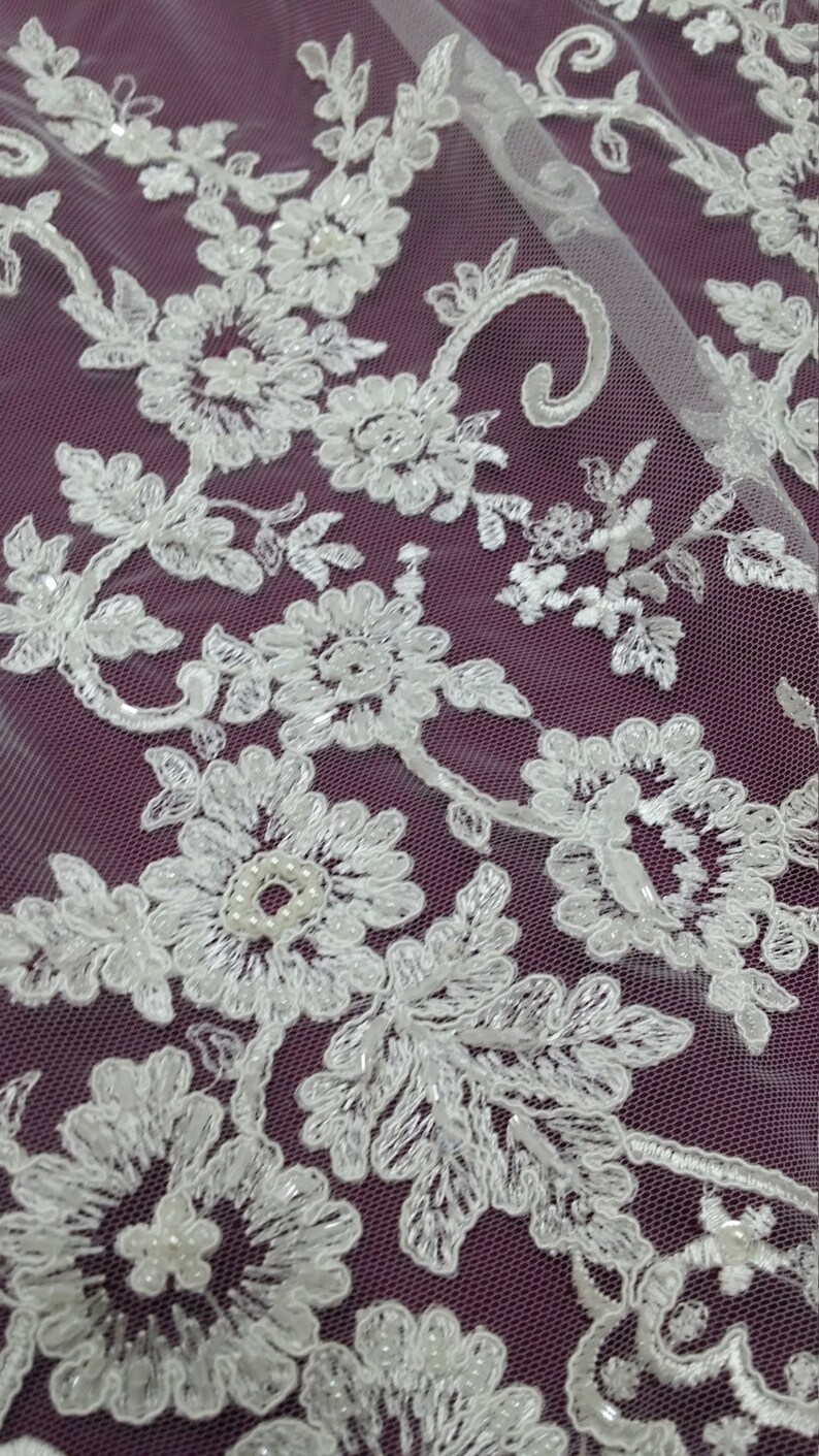 May include: Close-up of white floral lace fabric with intricate embroidered details. The lace features a delicate pattern of flowers, leaves, and beaded accents. The fabric is set against a deep purple background, creating a contrast that highlights the lace's design.