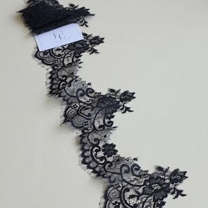 Könnte beinhalten: Ein Stück schwarze Spitzenborte mit einem komplizierten Blumenmuster und gewellten Kanten. Ein kleines weißes rechteckiges Etikett mit dem schwarzen Text "LT LACE TO LOVE" ist am oberen Teil des gefalteten schwarzen Spitzenstoffs befestigt.