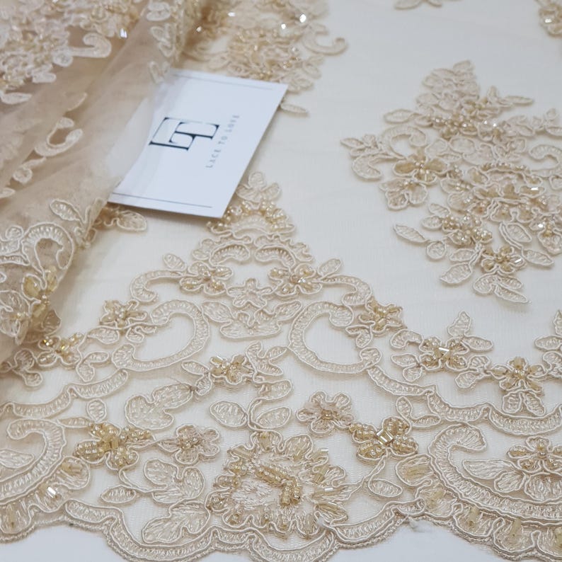 May include: Close-up of beige lace fabric featuring detailed floral and scrollwork patterns. The lace is adorned with tiny beads, giving it a delicate and luxurious texture. A tag with the text "LACE TO LOVE" is visible.