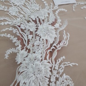 Puede incluir: Tela de encaje floral blanca con adornos de cuentas. El encaje presenta un delicado patr&oacute;n floral con detalles intrincados. La tela es transparente y ligera, lo que la hace ideal para una variedad de proyectos.