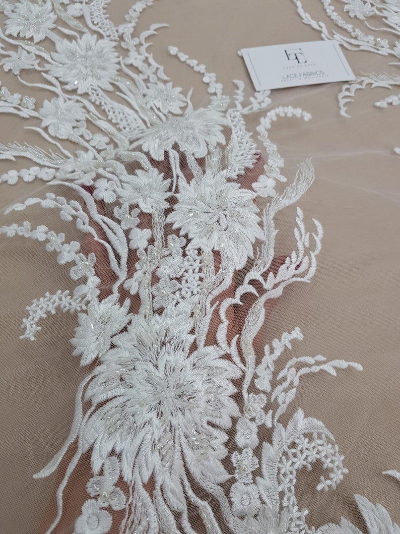 Puede incluir: Tela de encaje floral blanca con adornos de cuentas. El encaje presenta un delicado patr&oacute;n floral con detalles intrincados. La tela es transparente y ligera, lo que la hace ideal para una variedad de proyectos.