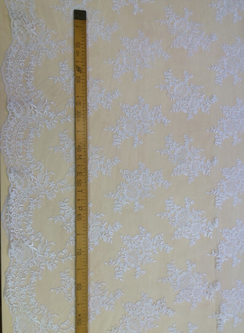 May include: White embroidered lace fabric with a delicate floral pattern. The fabric is shown with a ruler measuring 90 cm.