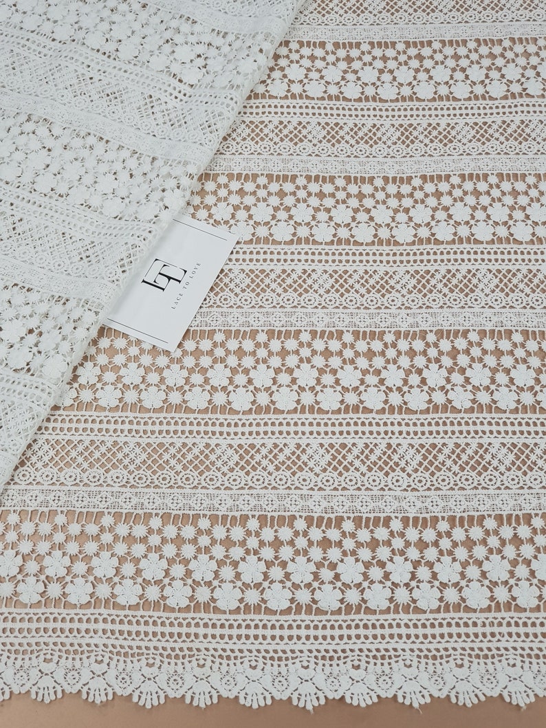 May include: White crochet lace fabric with a repeating pattern of flowers and geometric shapes. The lace has a scalloped edge.