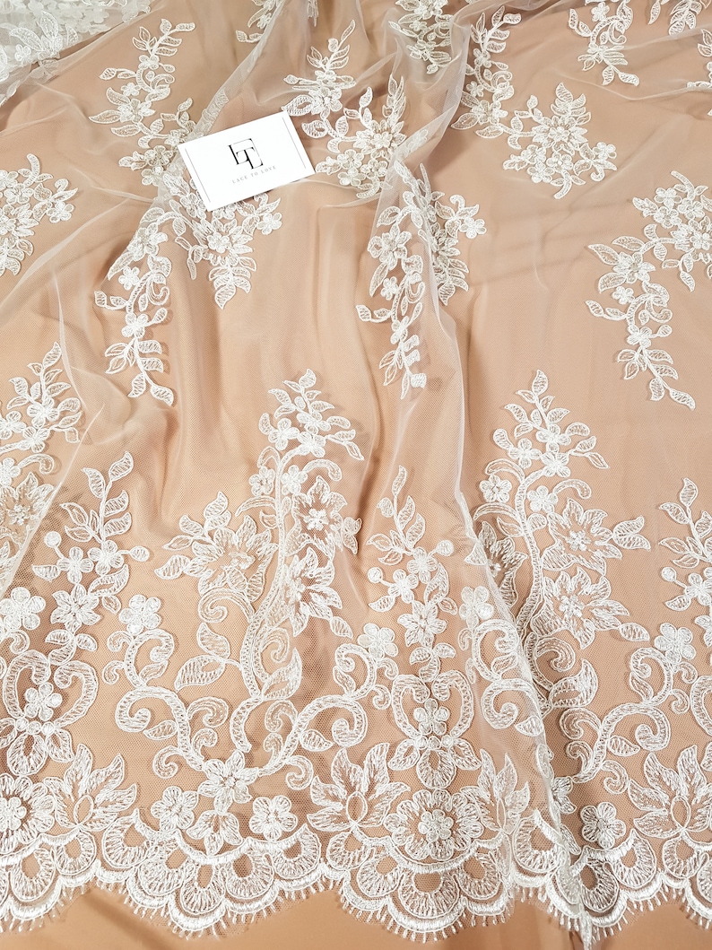 May include: White floral lace fabric with a delicate pattern. The lace is on a sheer, beige mesh background.
