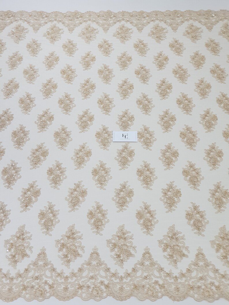 May include: Beige lace fabric with a repeating floral pattern and a scalloped edge. The delicate design features embroidered flowers and small embellishments, creating a textured surface. The fabric is suitable for various craft and design projects.