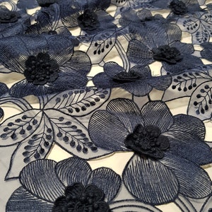 May include: A navy blue lace fabric featuring a repeating pattern of large embroidered flowers and delicate leaves. The design includes three-dimensional floral centres with intricate stitching and openwork details throughout the sheer mesh base.