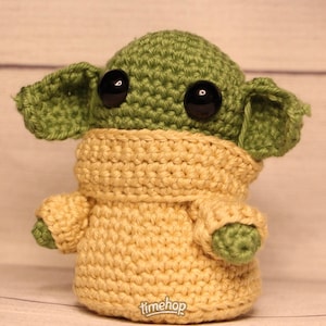May include: A crocheted green and beige plush toy with large black eyes. The toy is wearing a beige sweater.