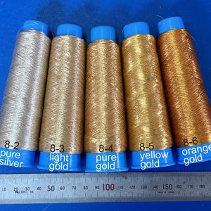Könnte beinhalten: Fünf Spulen mit Metallic-Garn in den Farben Silber, Hellgold, Reingold, Gelbgold und Orangegold. Die Spulen sind mit ihren entsprechenden Farben und Nummern 8-2, 8-3, 8-4, 8-5 und 8-6 beschriftet.