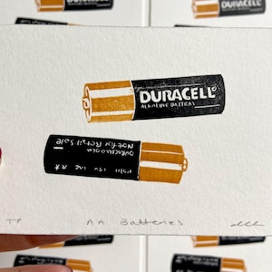 Puede incluir: Una impresi&oacute;n que muestra dos pilas AA Duracell, una en posici&oacute;n vertical y otra invertida. Las pilas son negras con detalles dorados y el logotipo de Duracell. El texto "AA Batteries" est&aacute; en la parte inferior.