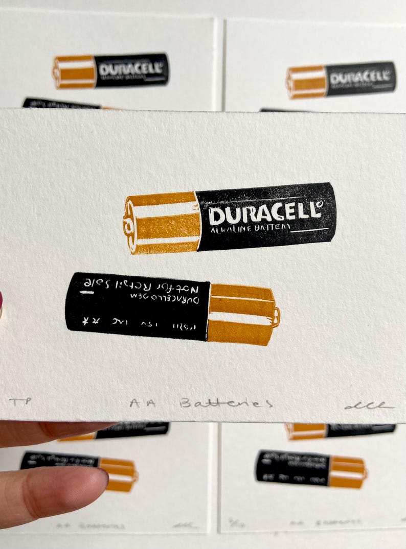 Puede incluir: Una impresi&oacute;n que muestra dos pilas AA Duracell, una en posici&oacute;n vertical y otra invertida. Las pilas son negras con detalles dorados y el logotipo de Duracell. El texto "AA Batteries" est&aacute; en la parte inferior.