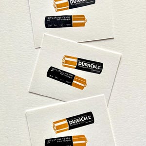 Puede incluir: Varias tarjetas blancas con una ilustraci&oacute;n en negro y dorado de una pila alcalina Duracell. El texto "DURACELL ALKALINE BATTERY" est&aacute; impreso en la pila. La pila inferior tiene el texto "Not for Retail Sale".