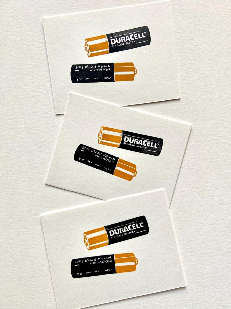 Puede incluir: Varias tarjetas blancas con una ilustraci&oacute;n en negro y dorado de una pila alcalina Duracell. El texto "DURACELL ALKALINE BATTERY" est&aacute; impreso en la pila. La pila inferior tiene el texto "Not for Retail Sale".