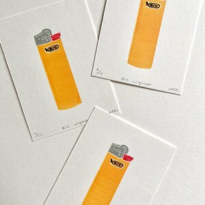 Op de afbeelding: Drie prints van een gele BIC aansteker met grijze en rode accenten op wit papier. De tekst "BIC Lighter" en een breuk van 15 staan onderaan elke print.