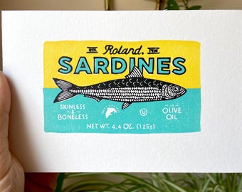 Pode incluir: Impressão de uma lata de sardinhas com o texto "Roland. SARDINES" em amarelo e azul. A lata apresenta uma ilustração de peixe a preto e branco e indica "Sem pele e sem espinhas" e "Azeite". Peso líquido 125g.