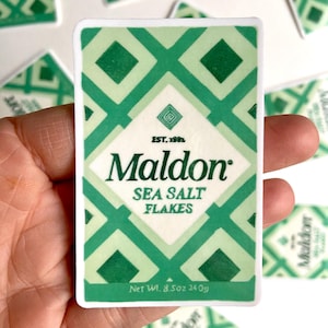 Może przedstawiać: Naklejka z zielono-białym geometrycznym wzorem, z napisem "Maldon Sea Salt Flakes". Naklejka zawiera tekst "Waga netto 240g". Marka "Maldon" jest pogrubiona, z rokiem "EST. 1982".