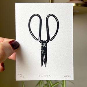 May include: A black and white linocut print of scissors on white paper. The word "scissors" is written in cursive below the image. The print is numbered 20/OE and signed "dee".