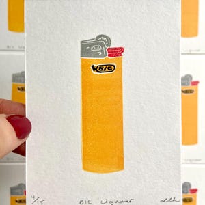 Op de afbeelding: Een print van een gele BIC aansteker met grijze en rode accenten. De tekst "BIC lighter" staat onder de afbeelding, samen met het editienummer 4/15. De aansteker staat tegen een witte achtergrond, met andere prints zichtbaar.