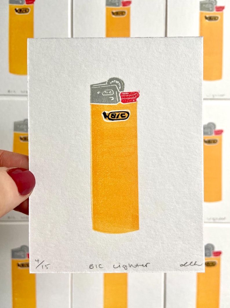 Op de afbeelding: Een print van een gele BIC aansteker met grijze en rode accenten. De tekst "BIC lighter" staat onder de afbeelding, samen met het editienummer 4/15. De aansteker staat tegen een witte achtergrond, met andere prints zichtbaar.