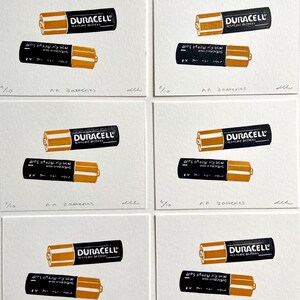Puede incluir: Una serie de seis grabados art&iacute;sticos con pilas AA Duracell. Cada grabado muestra dos pilas, una con el logotipo de Duracell y la otra con texto. Los grabados est&aacute;n numerados 3/10 y 7/10, con el texto "AA Batteries" debajo.