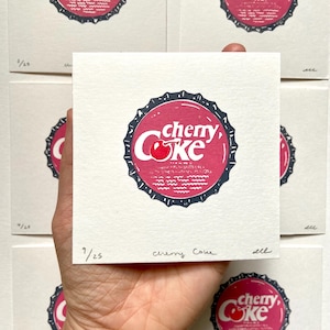 May include: A close-up of a red and white cherry Coke bottle cap print. The print is on a white card with the number 9/25 and the words "cherry Coke" written in black ink.