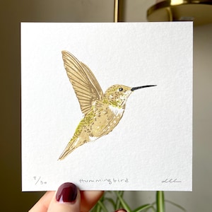 Op de afbeelding: Een vierkante print met een gedetailleerde illustratie van een kolibrie in vlucht. De vogel is afgebeeld in goud- en groentinten, met een lange zwarte snavel. De print is genummerd 8/30 en bevat het woord "Hummingbird".