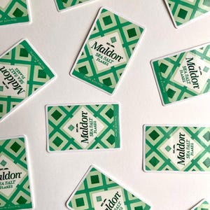 Może przedstawiać: Widok z g&oacute;ry przedstawia wiele prostokątnych pudełek Maldon Sea Salt Flakes rozrzuconych na białej powierzchni. Każde pudełko ma zielony wz&oacute;r w romby i nazwę marki w eleganckiej czcionce. Pudełka zawierają 240g płatk&oacute;w soli morskiej.