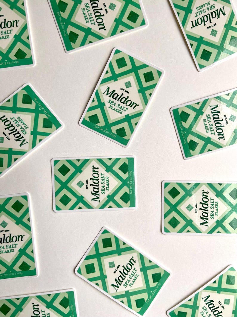Może przedstawiać: Widok z g&oacute;ry przedstawia wiele prostokątnych pudełek Maldon Sea Salt Flakes rozrzuconych na białej powierzchni. Każde pudełko ma zielony wz&oacute;r w romby i nazwę marki w eleganckiej czcionce. Pudełka zawierają 240g płatk&oacute;w soli morskiej.