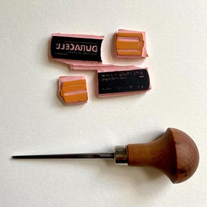 Puede incluir: Un conjunto de sellos de goma rosas con texto negro y detalles naranjas, junto con una herramienta de grabado de metal con mango de madera. Los sellos est&aacute;n fragmentados, mostrando la palabra "DURACELL" y otro texto. La herramienta de grabado tiene una punta.