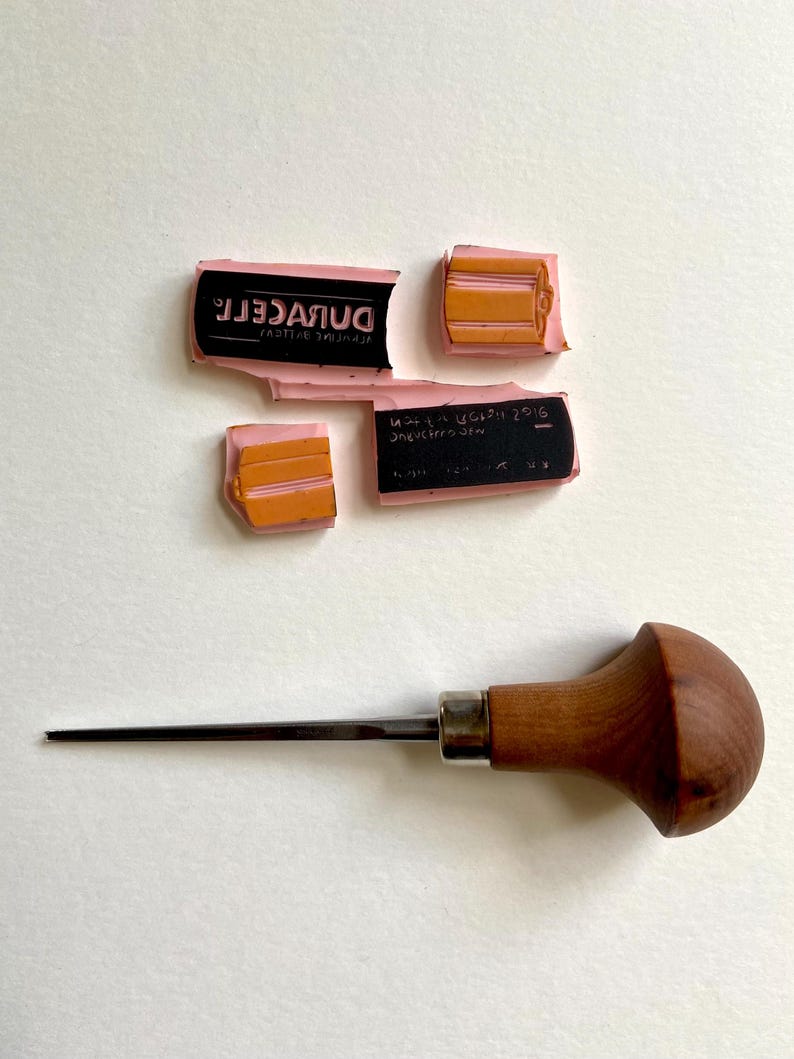 Puede incluir: Un conjunto de sellos de goma rosas con texto negro y detalles naranjas, junto con una herramienta de grabado de metal con mango de madera. Los sellos est&aacute;n fragmentados, mostrando la palabra "DURACELL" y otro texto. La herramienta de grabado tiene una punta.