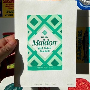 Może przedstawiać: Ręka trzyma wydruk pudełka Maldon Sea Salt Flakes z zielono-białym wzorem w romby. Tekst na pudełku brzmi "SEA SALT FLAKES" i "Net Wt. 240g". Inne wydruki związane z żywnością są w tle.