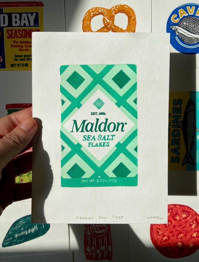 Może przedstawiać: Ręka trzyma wydruk pudełka Maldon Sea Salt Flakes z zielono-białym wzorem w romby. Tekst na pudełku brzmi "SEA SALT FLAKES" i "Net Wt. 240g". Inne wydruki związane z żywnością są w tle.