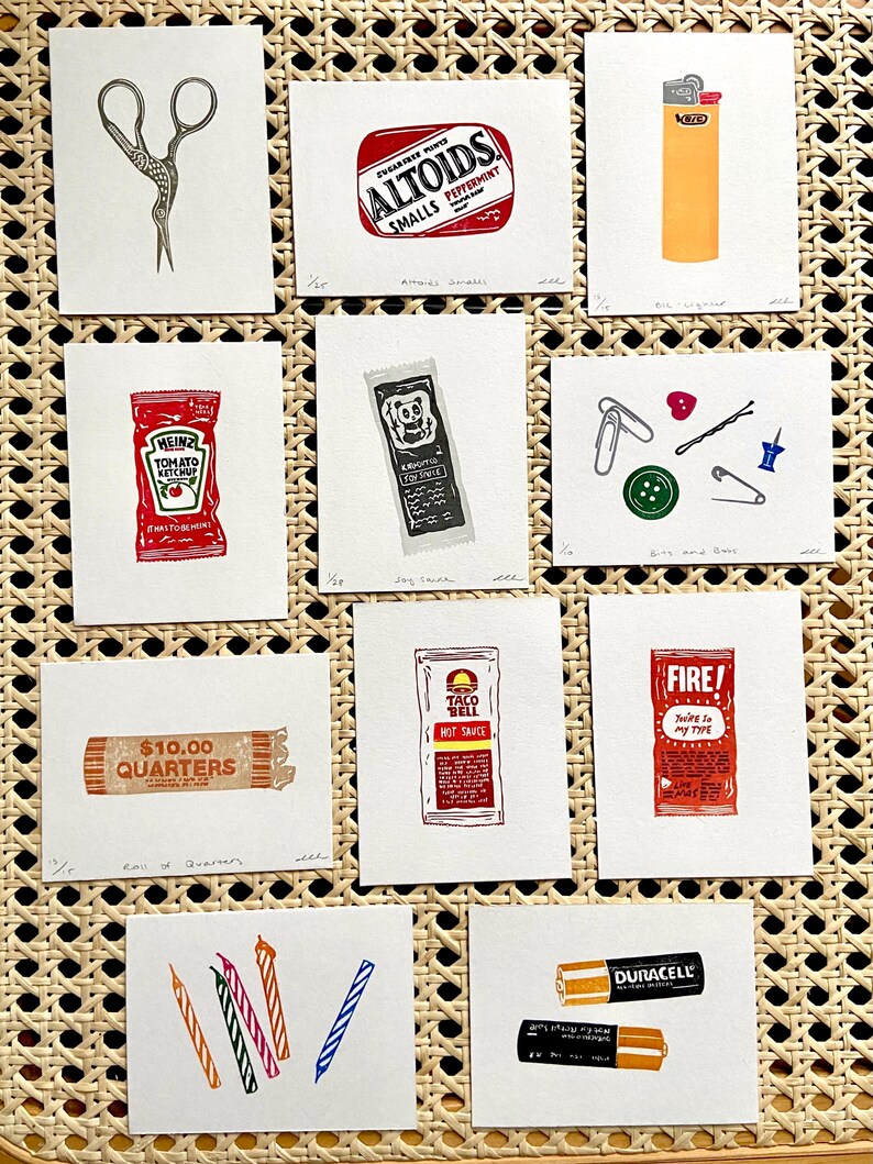 Op de afbeelding: Een verzameling kleine, vierkante kunstprints met alledaagse voorwerpen. Prints zijn onder meer een schaar, een pakje Altoids, een aansteker, een ketchupzakje, een hot saus zakje, een rol kwartjes en batterijen.