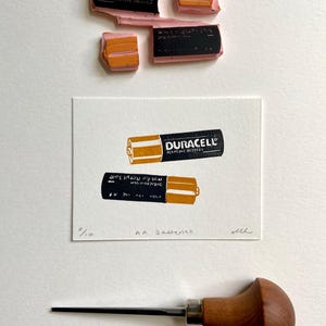 Puede incluir: Una impresi&oacute;n de dos pilas AA Duracell en tinta naranja y negra sobre papel blanco. La impresi&oacute;n est&aacute; etiquetada como "AA Batteries" y numerada "5/10". Tambi&eacute;n se ven sellos de tinta y una herramienta para tallar madera.