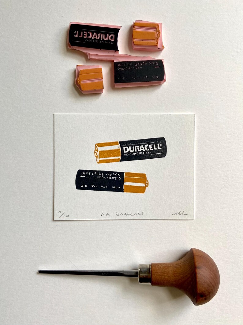 Puede incluir: Una impresi&oacute;n de dos pilas AA Duracell en tinta naranja y negra sobre papel blanco. La impresi&oacute;n est&aacute; etiquetada como "AA Batteries" y numerada "5/10". Tambi&eacute;n se ven sellos de tinta y una herramienta para tallar madera.