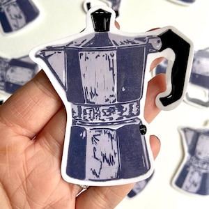 May include: A sticker featuring a blue and white illustration of a stovetop espresso maker. The sticker has a white border and a black handle. The design is a stylized drawing of a classic Italian coffee pot.