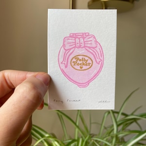 May include: A rectangular white card featuring a pink ink illustration of a heart-shaped Polly Pocket compact with a bow on top. The center of the compact is gold with the text Polly Pocket and a small heart. The card is signed Polly Pocket.