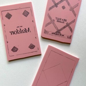 Może przedstawiać: Trzy r&oacute;żowe prostokątne bloki z czarnymi wzorami tuszu. Jeden blok ma słowo "Malden" odwr&oacute;cone, z tekstem "Est. 1897" nad nim. Inny blok brzmi "SEA SALT FLAKES" i "Net Wt 250g". Trzeci blok ma kształt rombu.