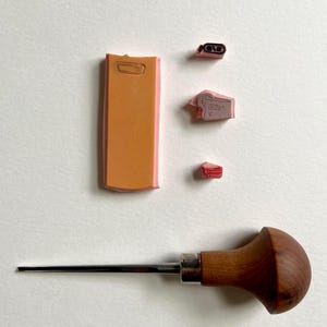 Op de afbeelding: Een assortiment knutselgereedschap en stempels. De afbeelding toont een houtbewerkingstool met houten handvat, verschillende roze en oranje rubberen stempels en een rechthoekige stempel met een handvat. De stempels zijn waarschijnlijk voor het maken van kleine ontwerpen of patronen.