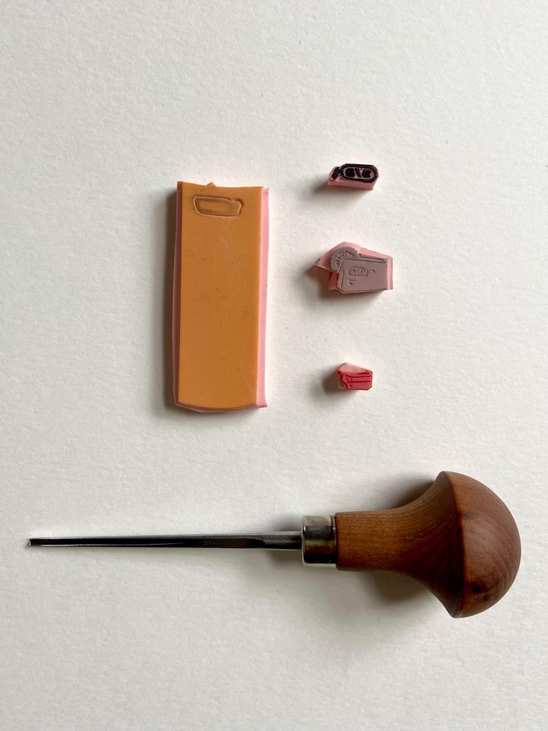 Op de afbeelding: Een assortiment knutselgereedschap en stempels. De afbeelding toont een houtbewerkingstool met houten handvat, verschillende roze en oranje rubberen stempels en een rechthoekige stempel met een handvat. De stempels zijn waarschijnlijk voor het maken van kleine ontwerpen of patronen.