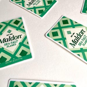Może przedstawiać: Kilka opakowań Maldon Sea Salt Flakes rozrzuconych na białej powierzchni. Każde opakowanie ma zielono-biały geometryczny wz&oacute;r, nazwę marki i szczeg&oacute;ły produktu. Waga netto wynosi 240g.